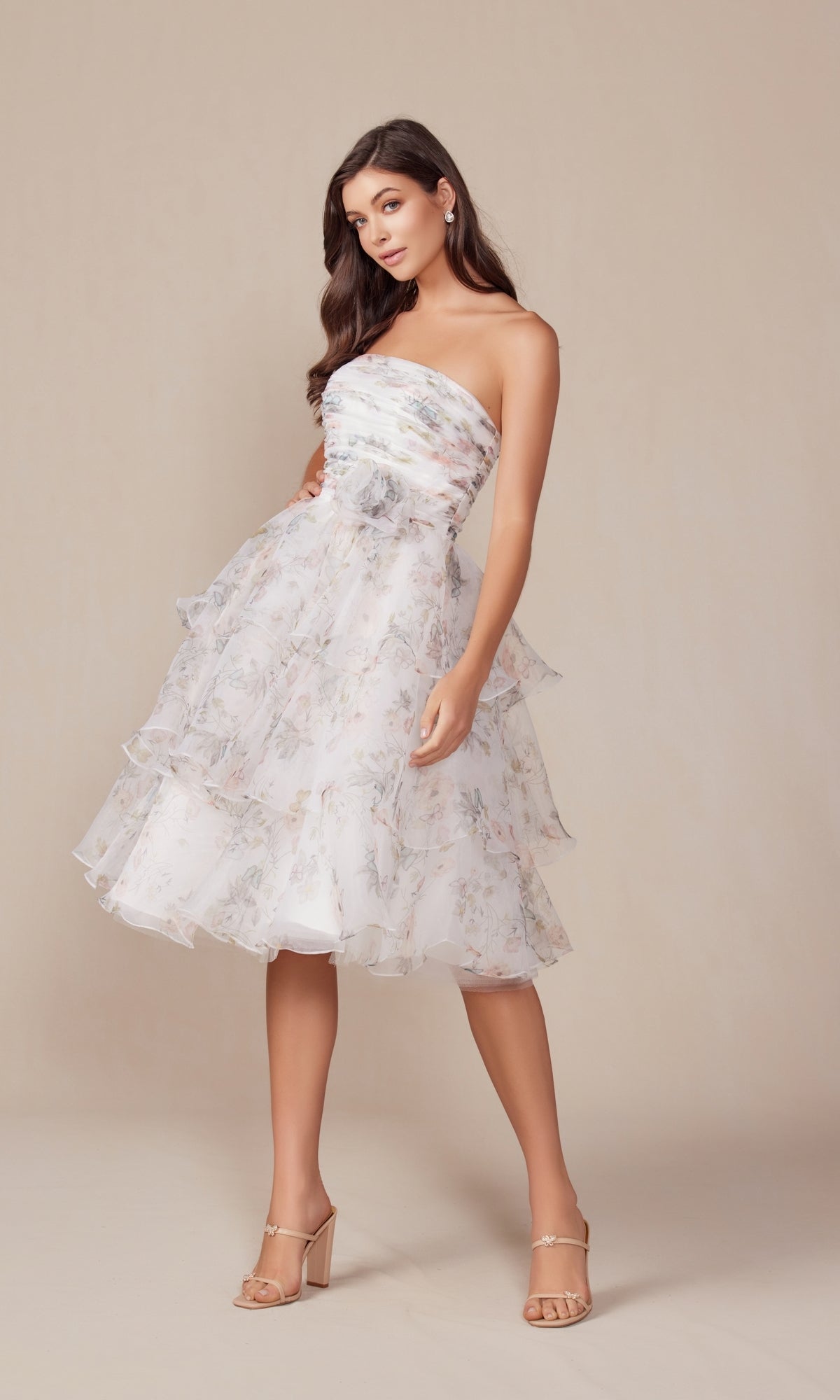 A woman wears the Nox Anabel Q842 Strapless Midi Print Hoco Dress, featuring a floral print and tiered layers, paired with nude high-heeled sandals, standing against a neutral background.
